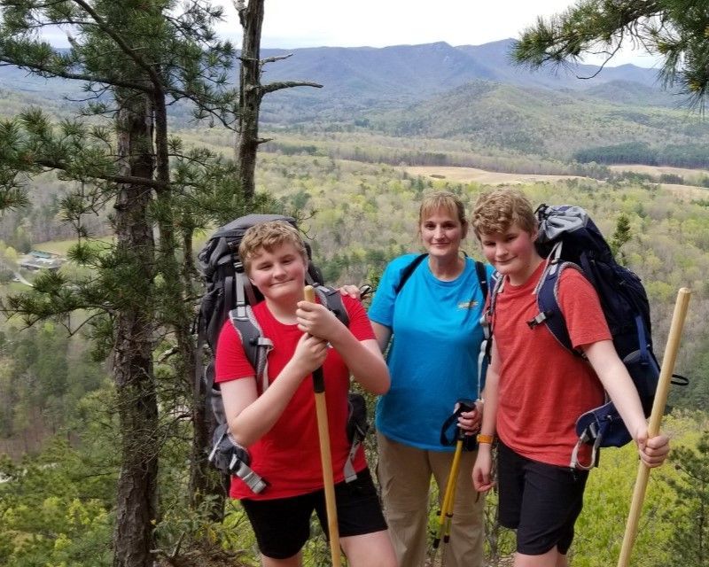 Three hikers with backpacks posing on a mountain trail with a scenic valley and hills behind them
