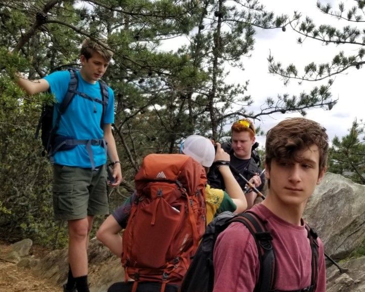 Hikers with backpacks resting on a rocky trail under pine trees, one person in foreground looking at camera