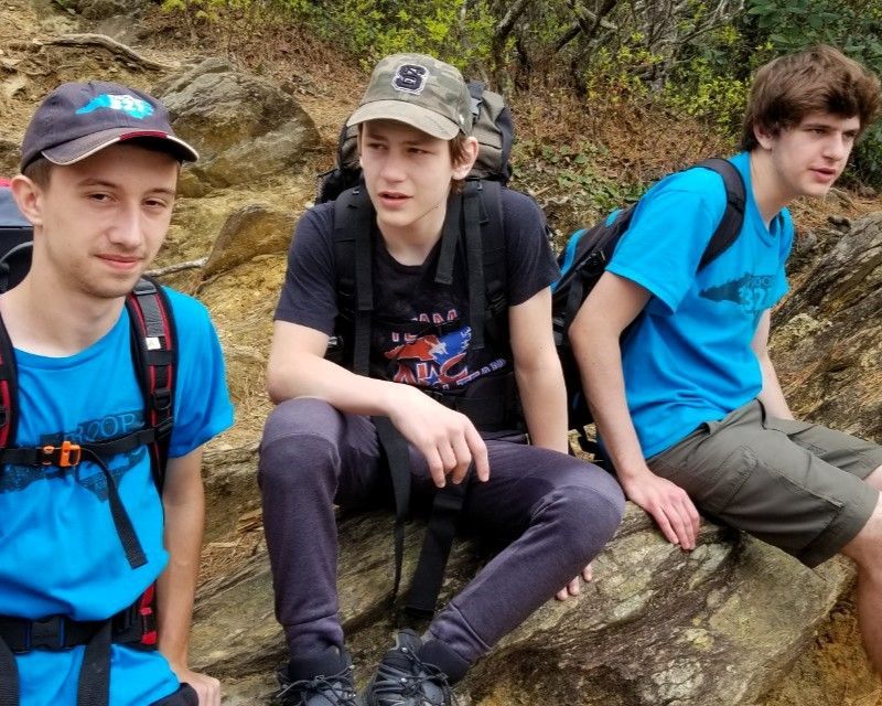 Three hikers in blue shirts resting on rocks in a wooded area, one wearing a cap and backpack.