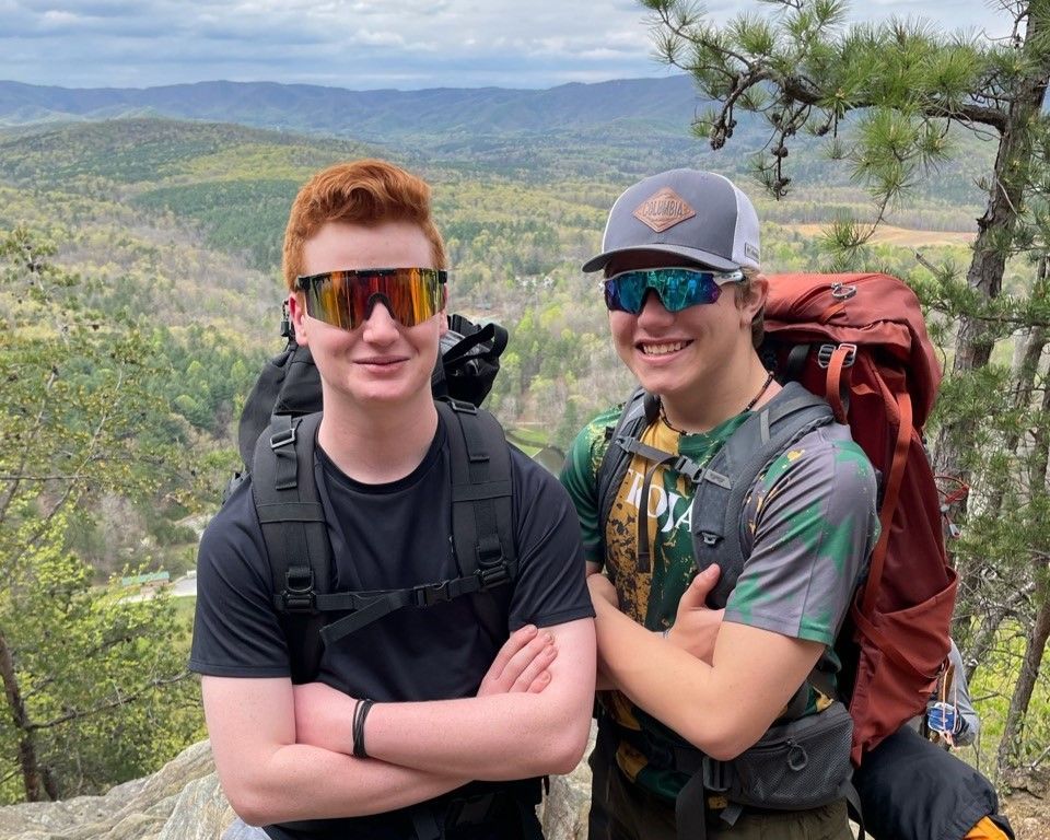 Two hikers smiling on a mountain overlook, wearing backpacks and sunglasses with a forested valley behind them