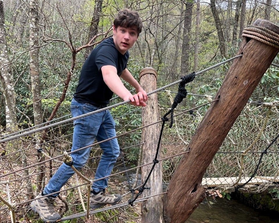 Person in black shirt and jeans crossing a rope bridge in a wooded area.