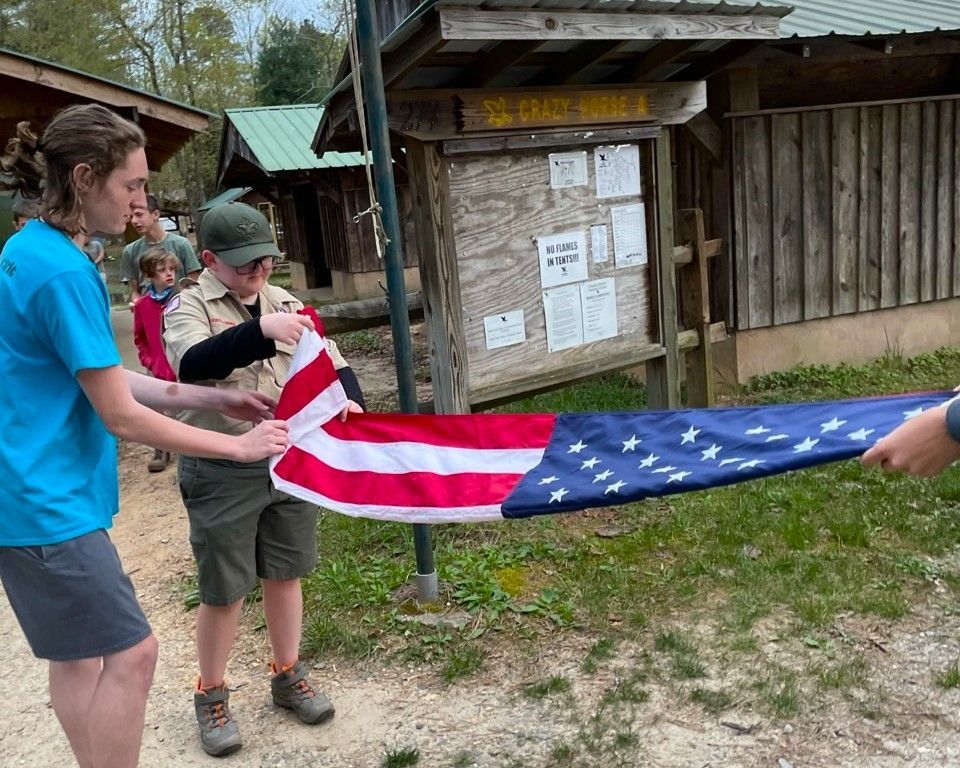 Two people hold a large American flag outdoors near a wooden bulletin board.