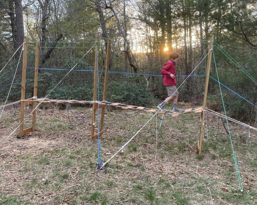 Person in a red jacket walking across a rope obstacle in a wooded area at sunset