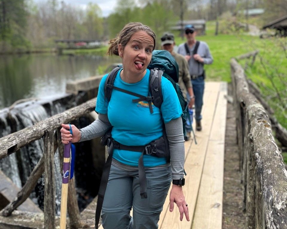 Hiker in blue shirt crossing a wooden bridge over water, smiling and carrying a backpack and trekking poles
