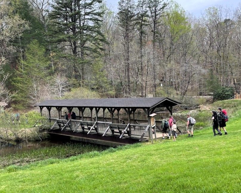 Covered wooden footbridge over a creek with hikers walking beside it in a grassy wooded park