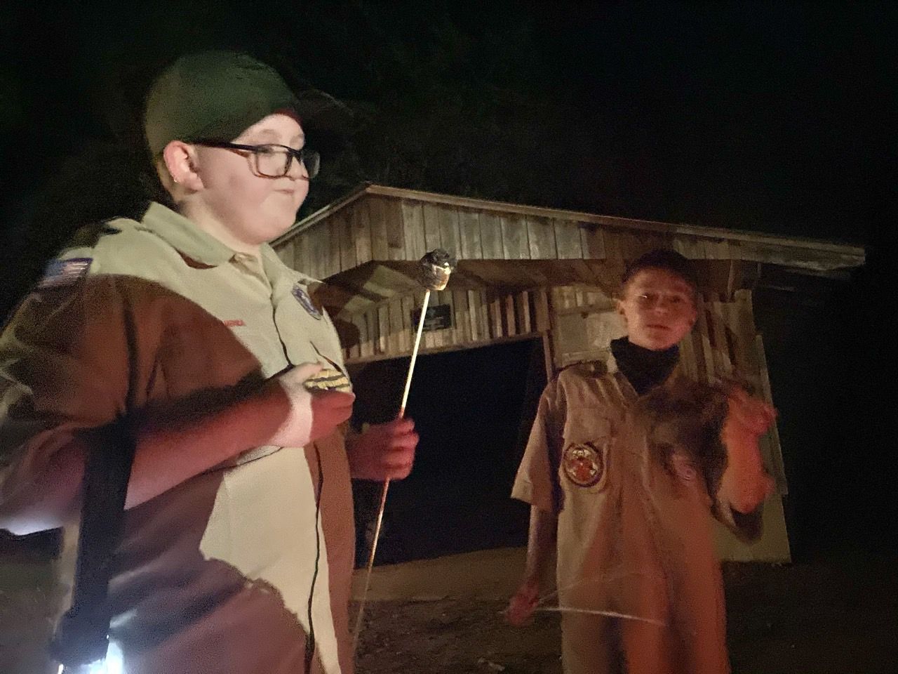 Two people holding marshmallow skewers near a lit outdoor fire at night