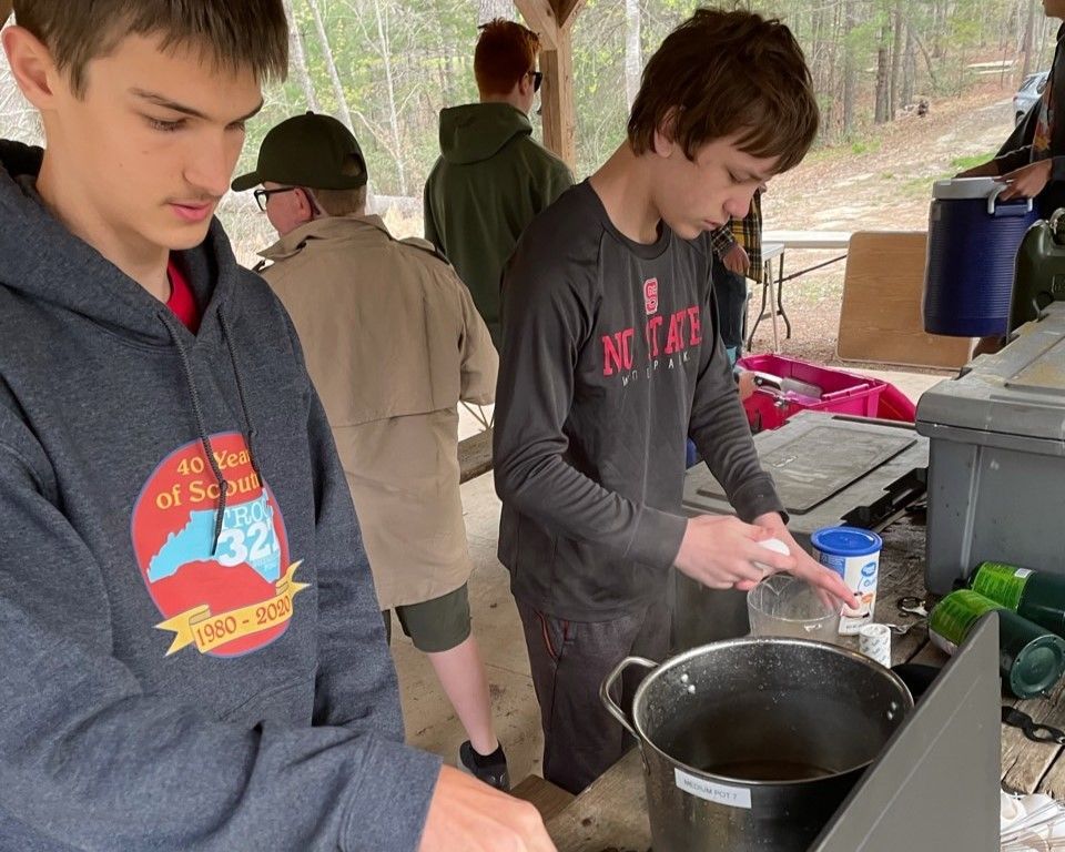 Two people cooking at an outdoor campsite, stirring a pot near a picnic table.