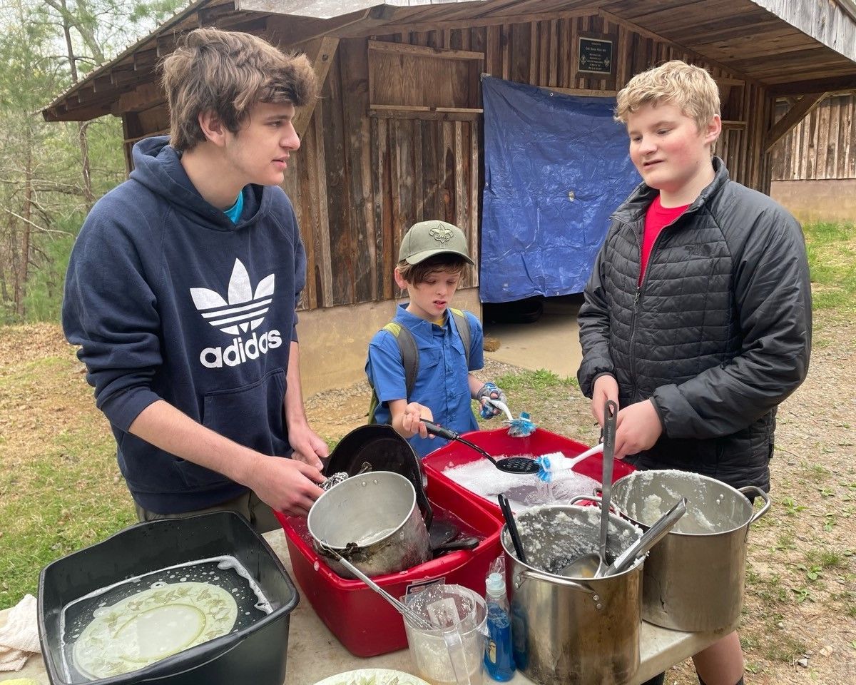 Three people stand outdoors by cooking pots and buckets in front of a rustic wooden cabin.