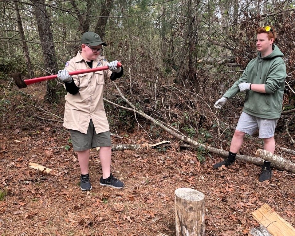 Two people outdoors carrying a red-handled saw and a log near a wooded trail