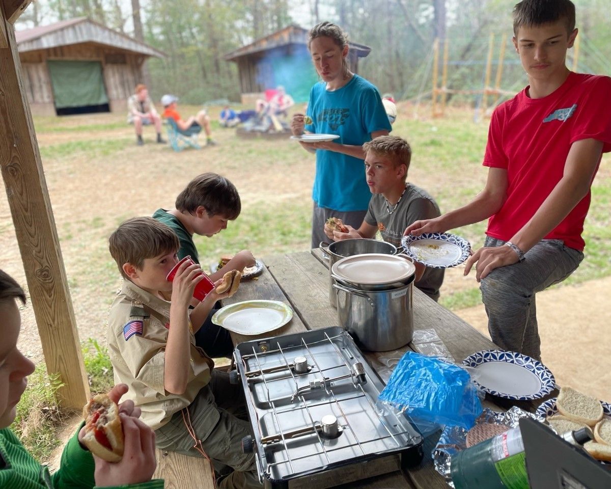 Children serving food at an outdoor buffet table with pots, trays, and disposable plates