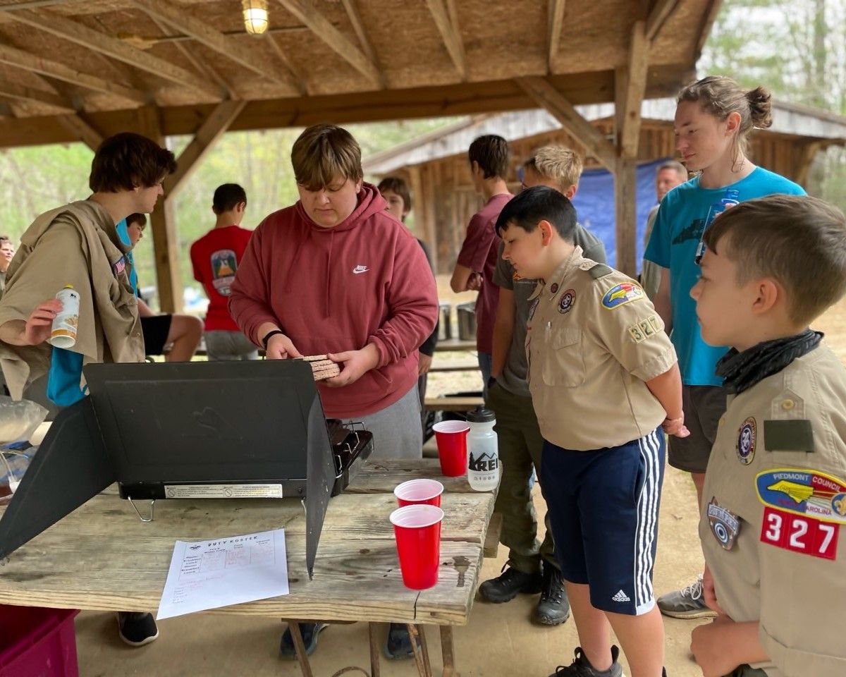 Scouts gathered around a laptop at a picnic table under a wooden pavilion, using cups and papers.