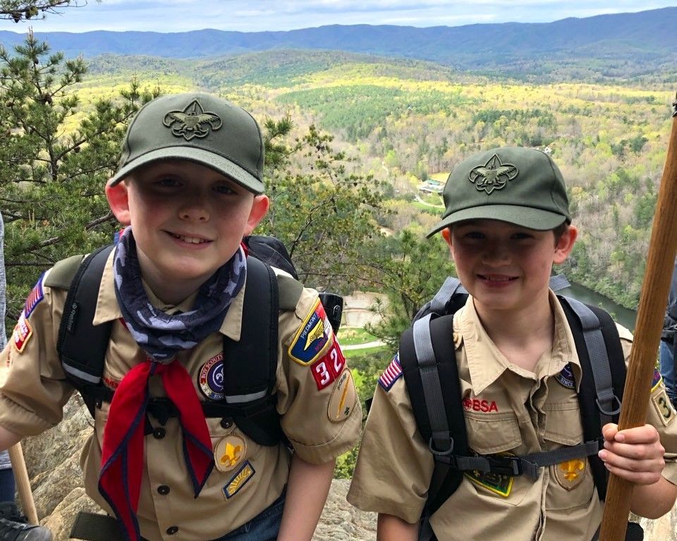 Two scouts in tan uniforms and green caps smiling on a mountain overlook with forested hills behind them