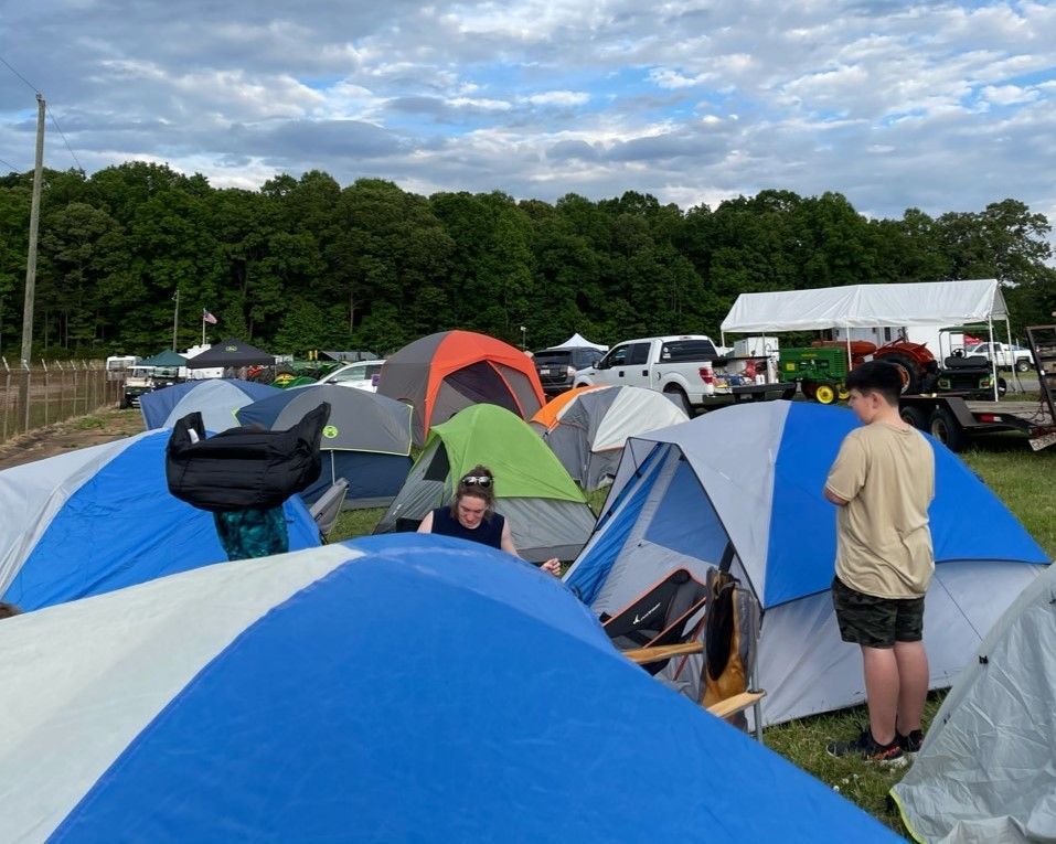 Campsite with colorful tents, parked vehicles, and people gathered outdoors under a cloudy sky.