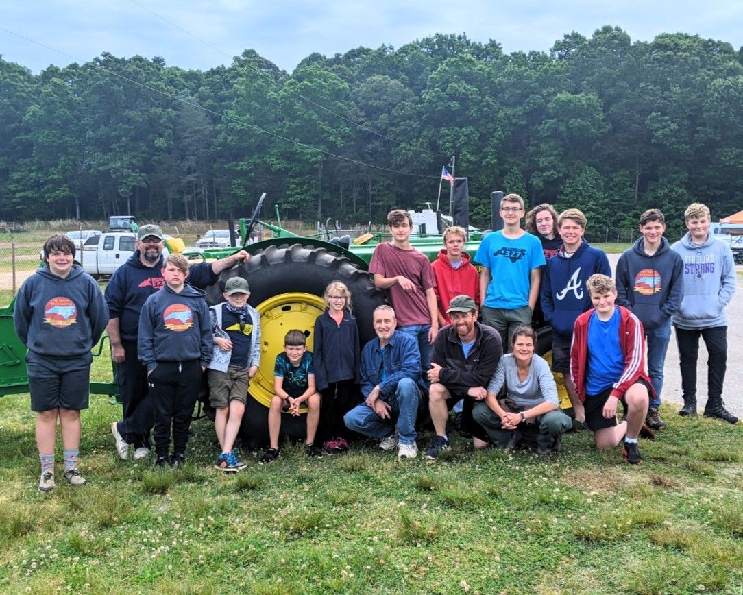Group of people posing outdoors in front of a green tractor on a grassy field.