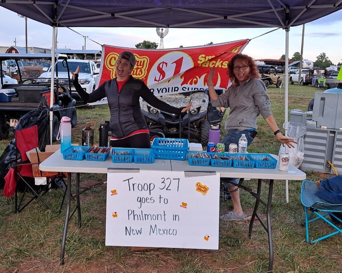 Two people at an outdoor market table with blue trays under a canopy, selling small items beside a handwritten sign.