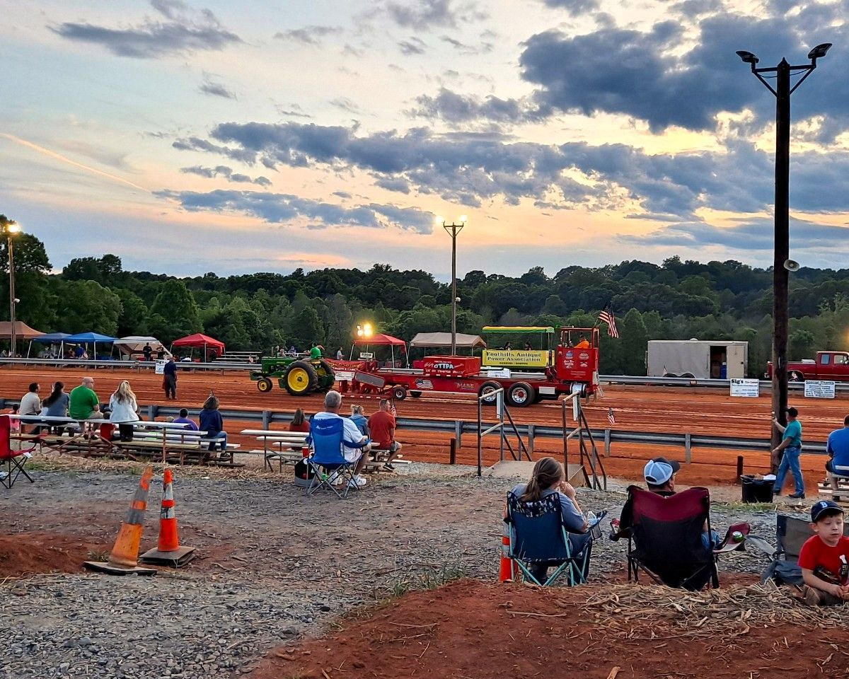 Dirt track tractor pull at dusk with spectators seated in folding chairs and orange traffic cones in the foreground