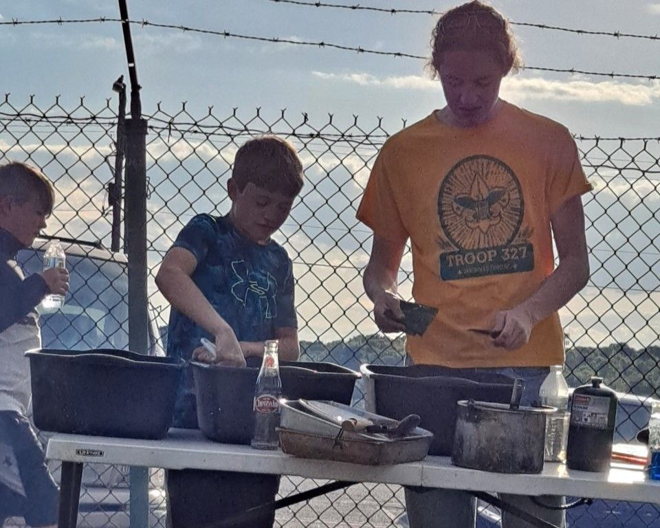 Three people cooking at a table outdoors by a chain-link fence at sunset