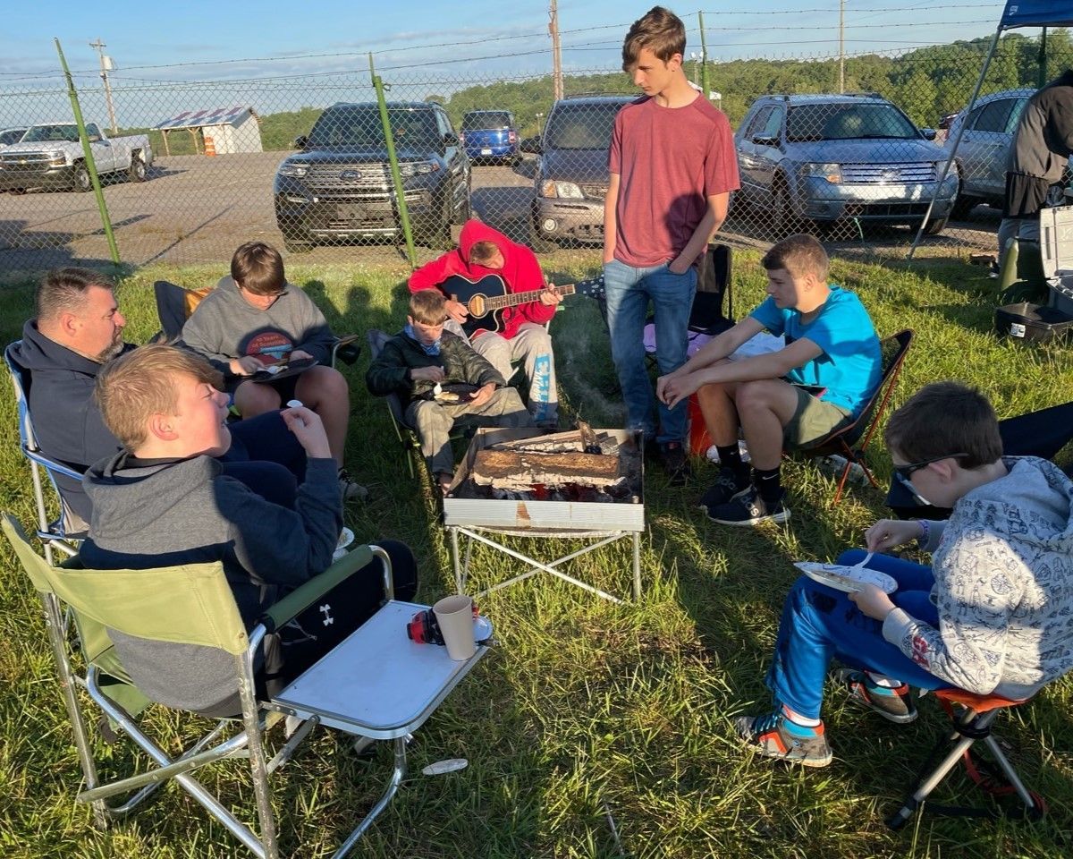 A group sits around a fire pit in folding chairs on a grassy campsite with parked cars nearby.