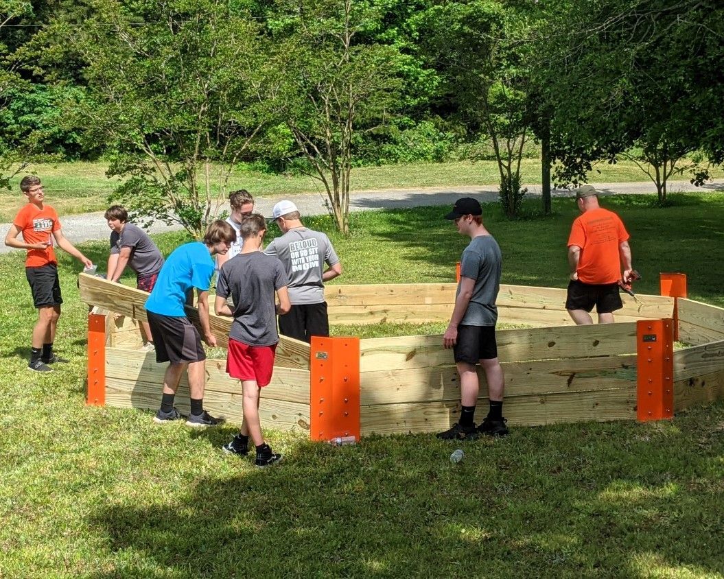 People assembling a wooden structure in a grassy park, with orange posts and several workers around it