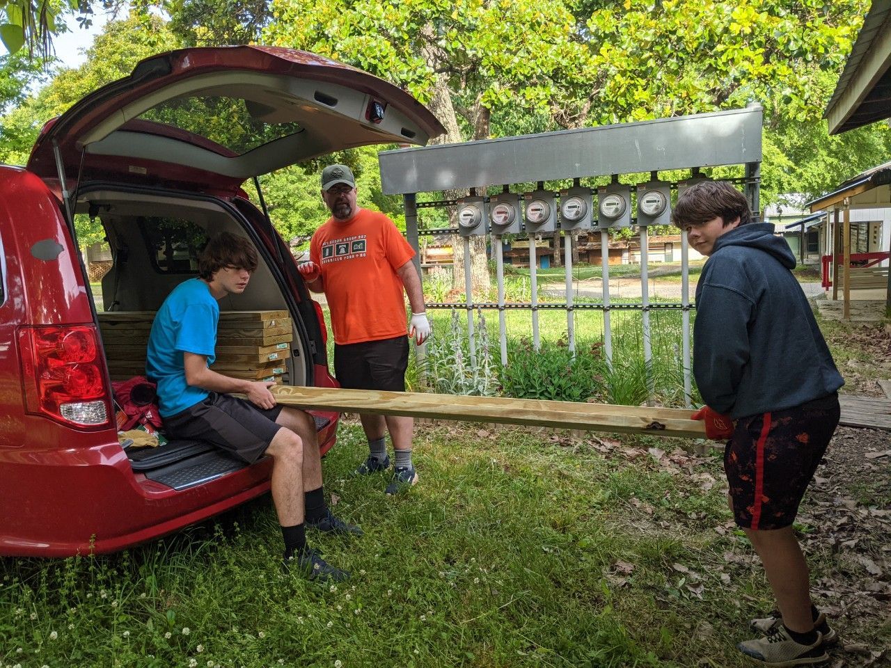 Three people loading a long wooden board from a red van in a grassy outdoor area