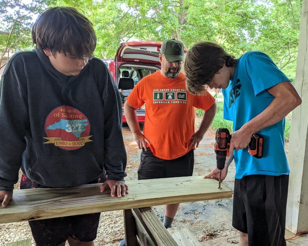 Three people work on a wooden board outdoors, using a power tool on a picnic table.
