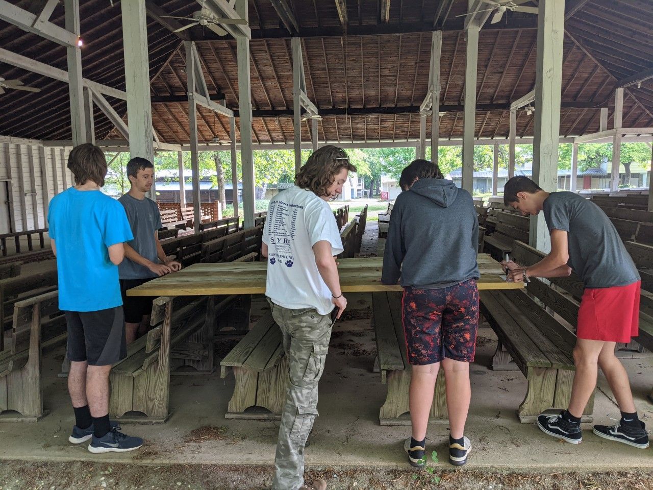 Five people stand around a long table in a rustic open-air shelter, working with trays and materials.