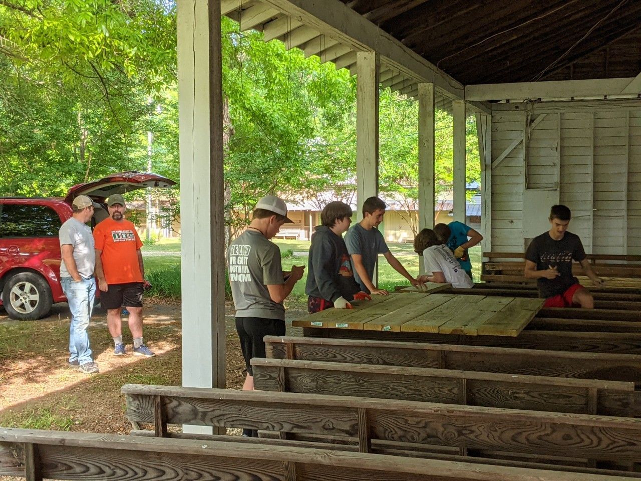 People gathered in a covered pavilion, seated and standing around wooden benches near a red SUV.