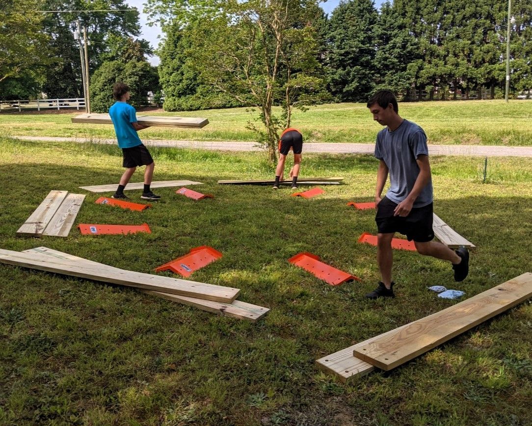 People playing cornhole in a sunny park, with red boards and a wooden walkway on the grass.