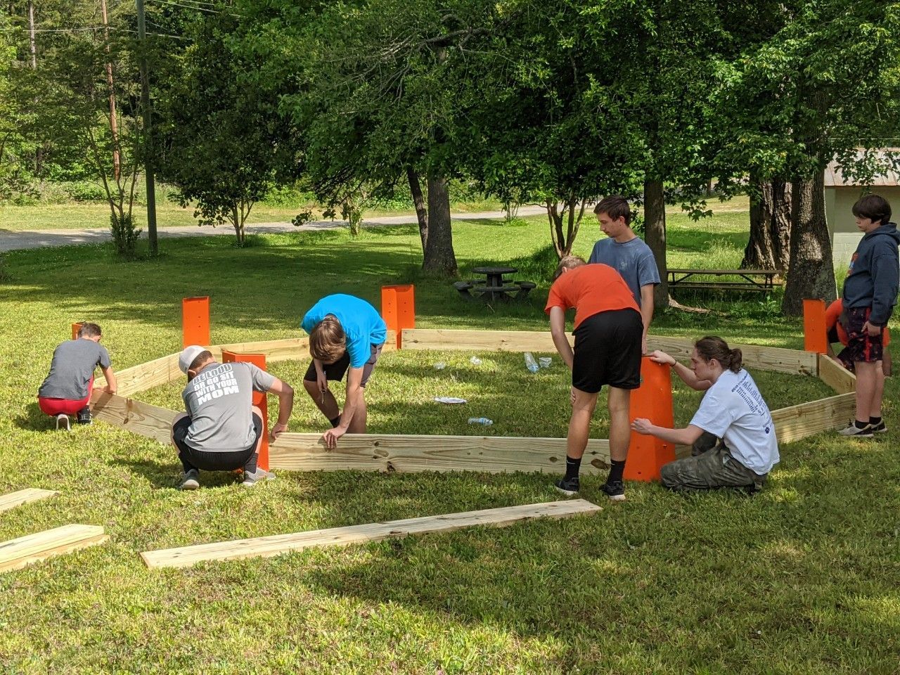People assembling a wooden frame outdoors in a grassy park, surrounded by trees and orange markers.