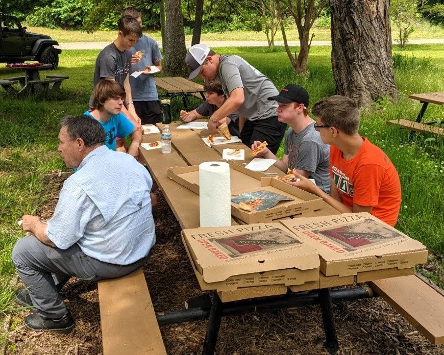People sharing pizza at a picnic table in a park, with a woman serving drinks nearby.