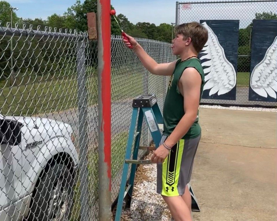 Person painting a red fence post beside a chain-link fence and ladder in a sunny outdoor lot
