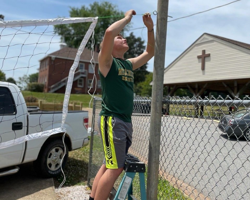 Person on a step stool hanging netting on a backyard soccer goal beside a parked truck and house