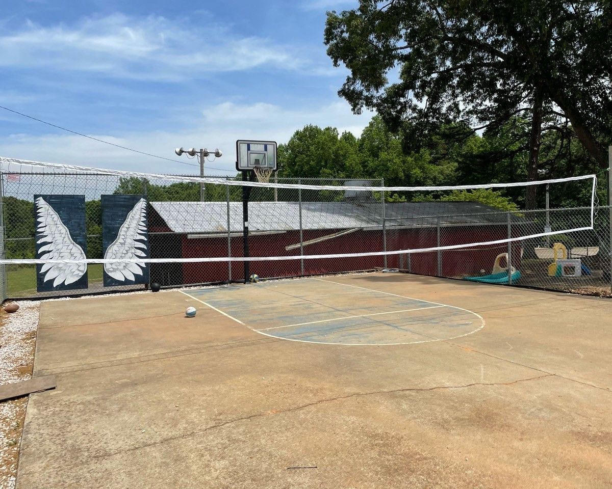 Outdoor volleyball court with a net, painted wings on the wall, and a cloudy sky beyond the fence