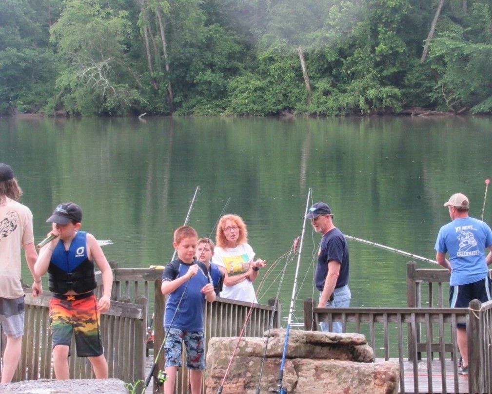 People fishing on a wooden dock by a calm lake, with trees reflected in the water.