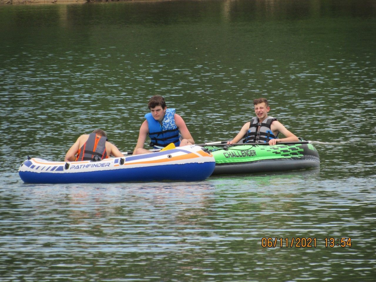 Three people on inflatable rafts floating on calm water, with a wooded shoreline in the background.