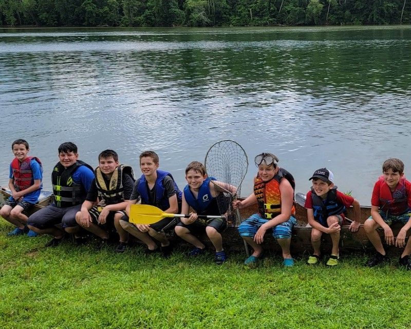 Group of children sitting by a riverbank, some holding paddles, with grassy shore and calm water behind them.