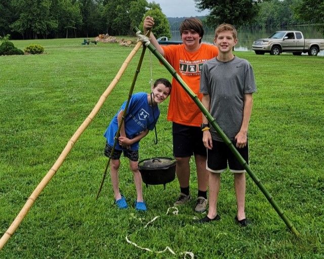 Three people posing by a campsite tripod with a hanging black pot in a grassy field