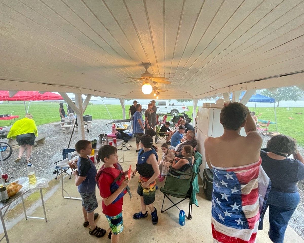 Children gather under a picnic shelter during a rainy outdoor party, with a table of snacks nearby.