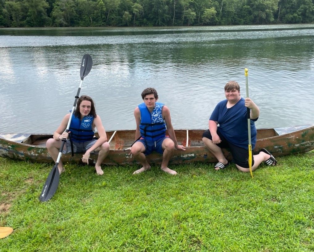 Three people in blue life vests sit by a lake with paddles, smiling on a grassy shore.