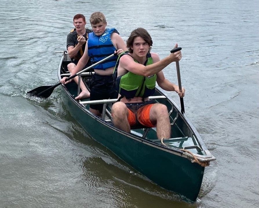 Three people paddle a green canoe on calm water, with one person in front steering.