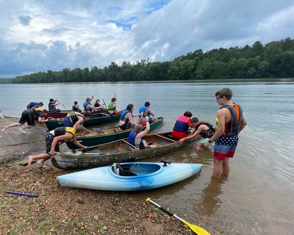 People launching kayaks into a river beside a pebbled shore under a cloudy sky