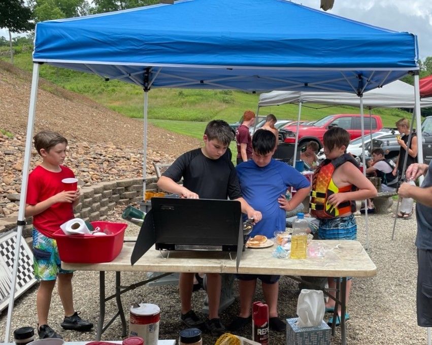 People serving food under a blue canopy at an outdoor picnic table, with drinks and snacks on display.