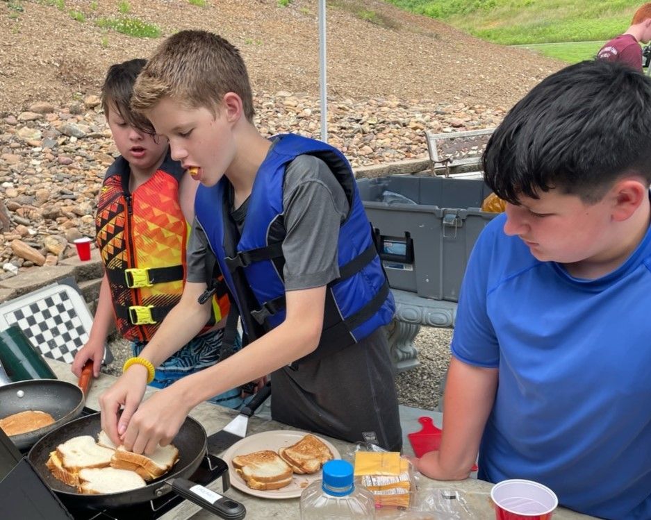 Three children cooking at an outdoor grill with burgers and bread on a picnic table