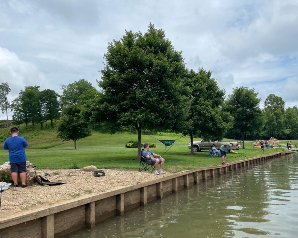 People fishing from a grassy shoreline beside a calm lake under cloudy skies.