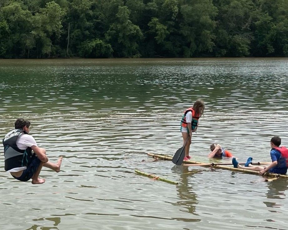 People on a small raft in a calm river, with trees lining the far bank.