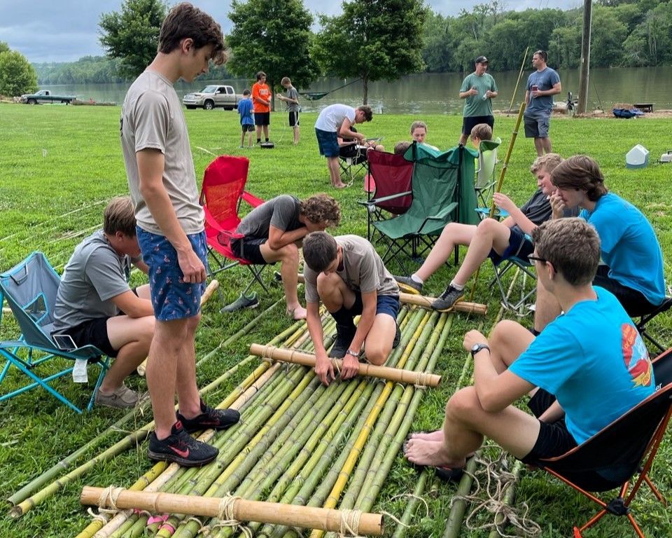 People building a bamboo raft on grass near a lake, with others watching and helping nearby