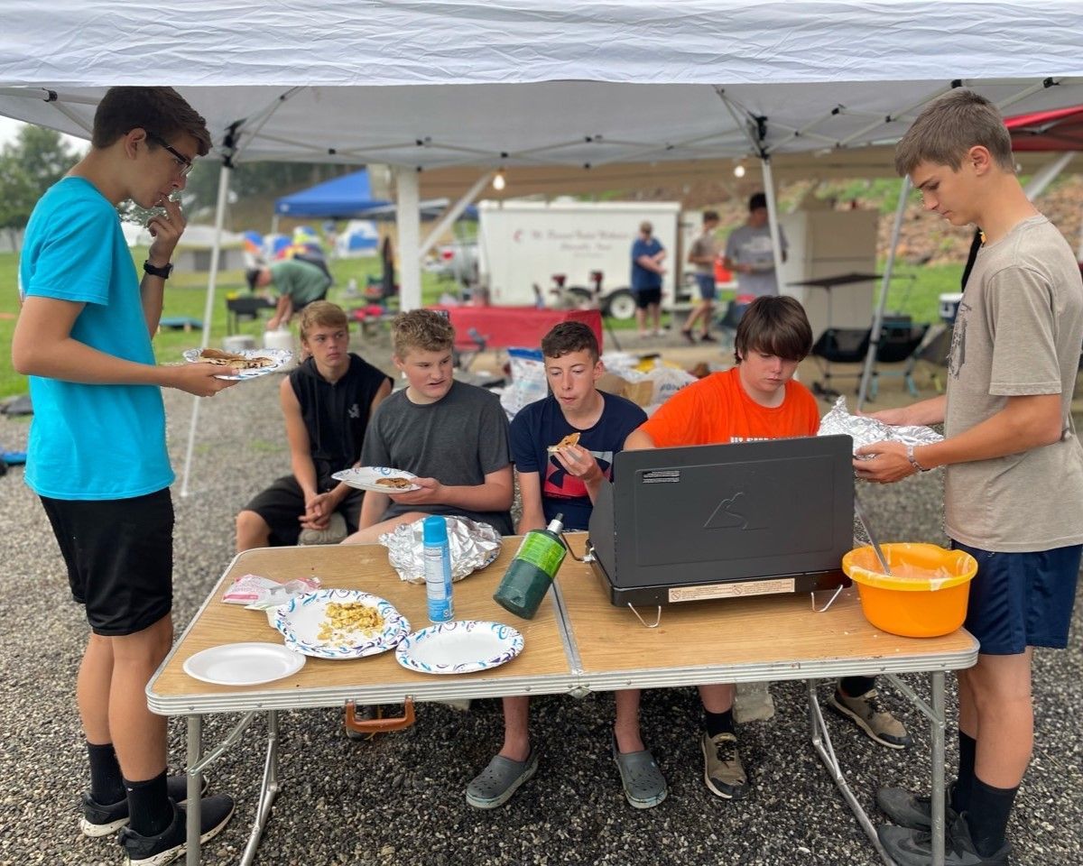 People gathered under a tent around a table with food, drinks, and a laptop at an outdoor event