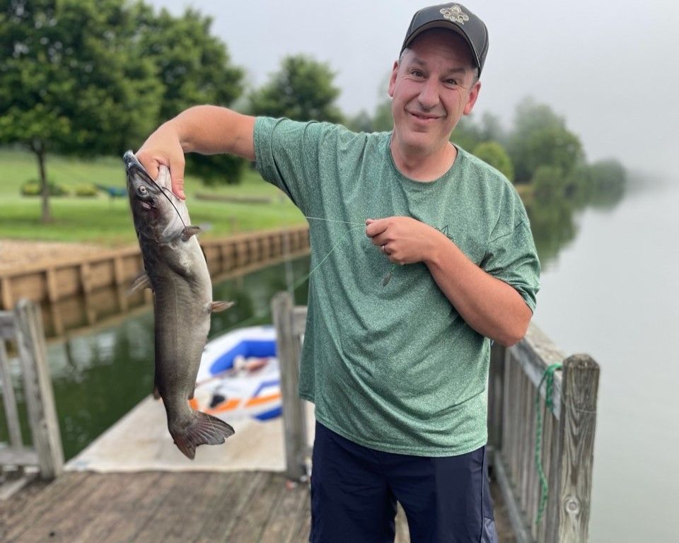 Man on a dock holding a large fish, with a misty lake and boat in the background.