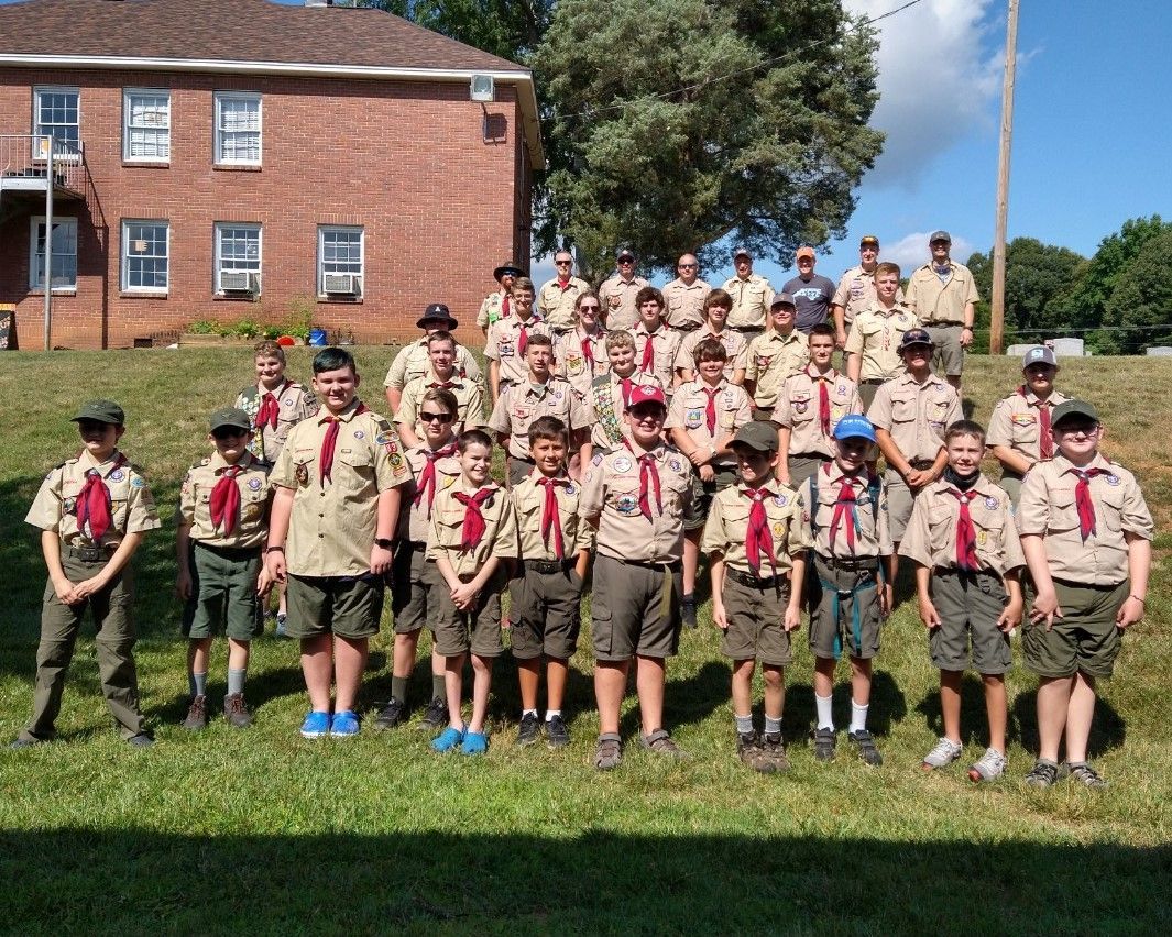 Group of scouts in tan uniforms and red neckerchiefs posing on a grassy field in front of a brick building.