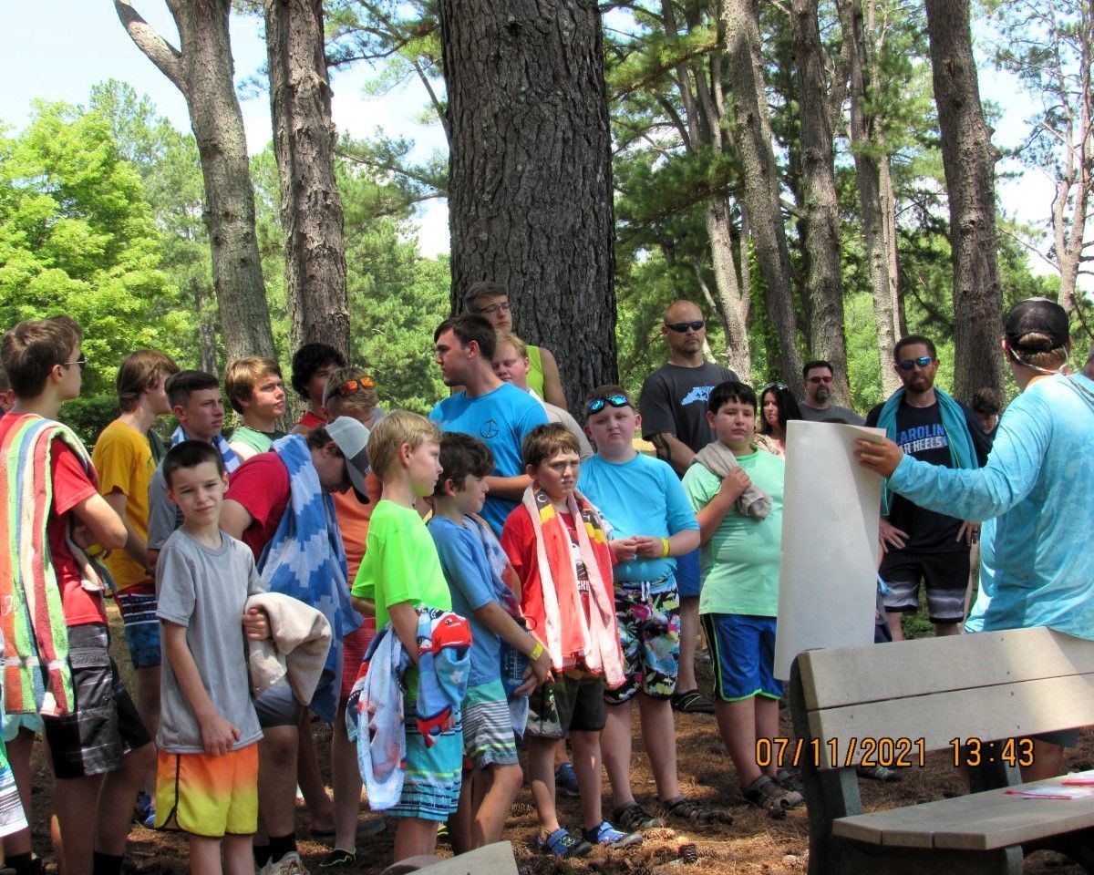 Children gathered outdoors listening to a speaker beside a picnic table in a wooded park.